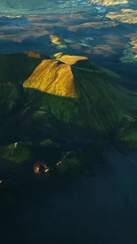 Golden hour drone aerial of a sunlit volcanic ridge and crater slopes, with long shadows stretching across vast highlands below. Epic Iceland landscape texture and depth.