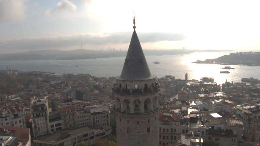 Stunning drone footage of the historic Galata Tower in Istanbul, Turkey, captured with panoramic views of the Bosphorus Strait and the city skyline. Boats move across the water while mosques and histo