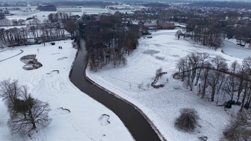 Aerial winter view of a winding river flowing through snowy fields and forests, with scattered houses and a distant town creating a calm, expansive winter landscape.