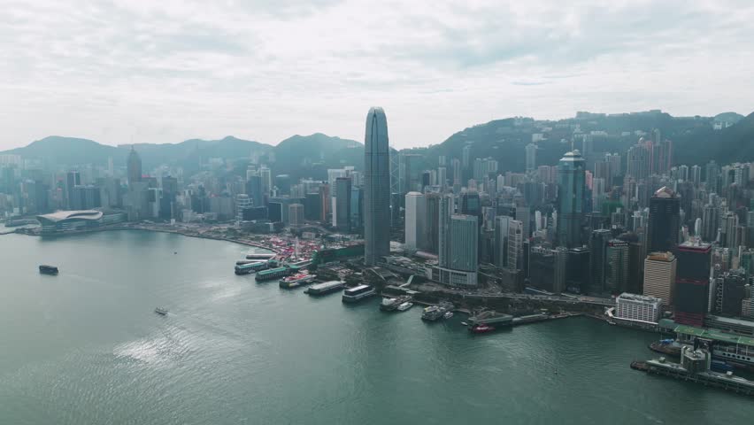 Panoramic aerial view of the skyscrapers and Victoria Harbour in downtown Hong Kong on a sunny day, showcasing the iconic skyline and waterfront.