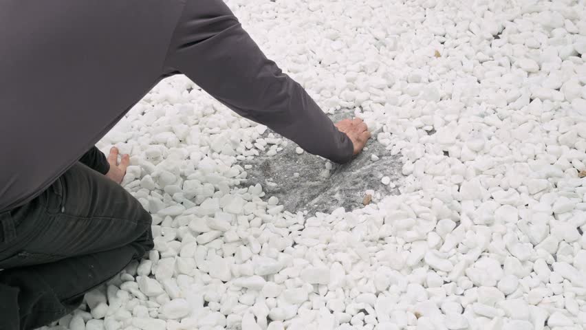 High angle shot of a person kneeling and using their hands to push aside white decorative pebbles in a backyard, creating a circular space on the ground for landscaping.