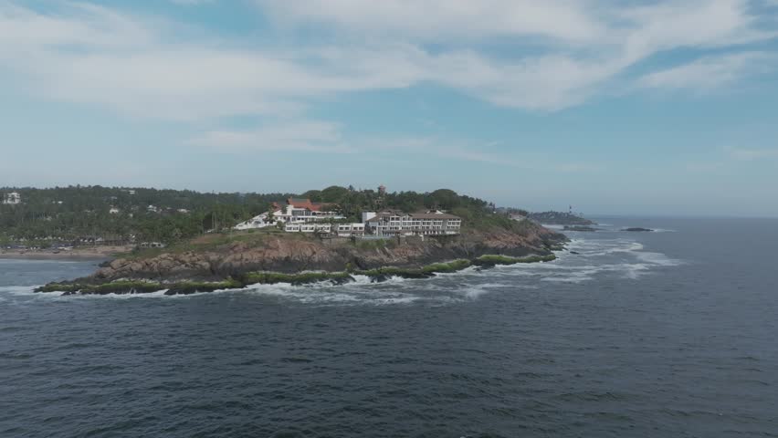 Aerial view of The Leela Kovalam resort perched on rocky cliffs, featuring tiered white buildings, palm accents, crashing Arabian Sea waves below under partly cloudy skies.