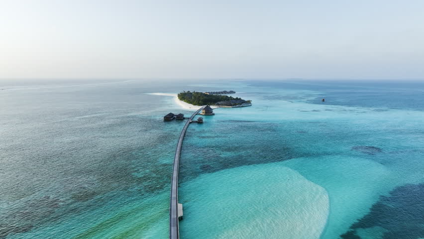 Aerial drone push in shot following a wooden walkway over the clear ocean leading to a lush green tropical island with overwater villas in the Maldives at sunrise
