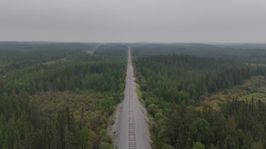 Aerial dolly-in flight over raised train tracks through boreal forest near Gilliam, Manitoba, Canada. Gravel and granite rail bed visible under a cloudy, smoky sky in cinematic northern wilderness.