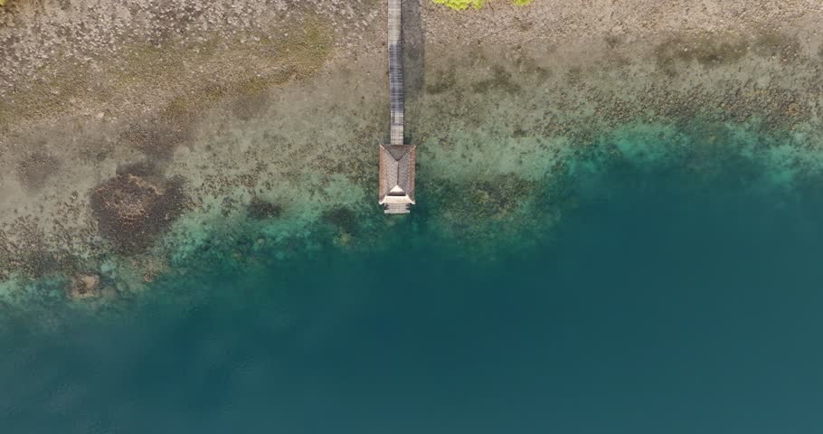 Top-down drone view of a small dock extending over turquoise lagoon and exposed coral reef in West Bali National Park, revealing marine textures, coastal patterns, and tropical island landscape
