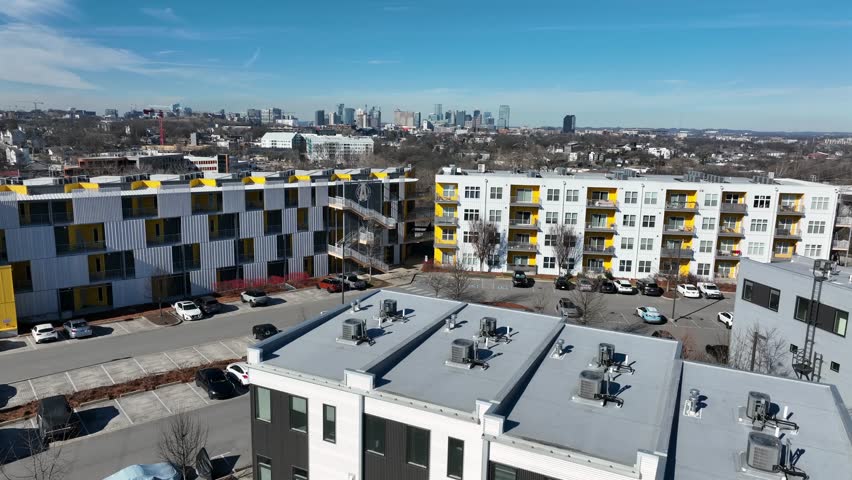 Aerial view of modern apartment buildings in quiet Nashville residential neighborhood with downtown skyline rising in distance under clear early spring sunlight. Wide shot. Rising shot. Early spring.