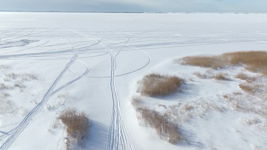 High-angle drone footage showing a vast expanse of a frozen lake covered in snow, marked with intricate intersecting patterns of snowmobile and vehicle tracks.