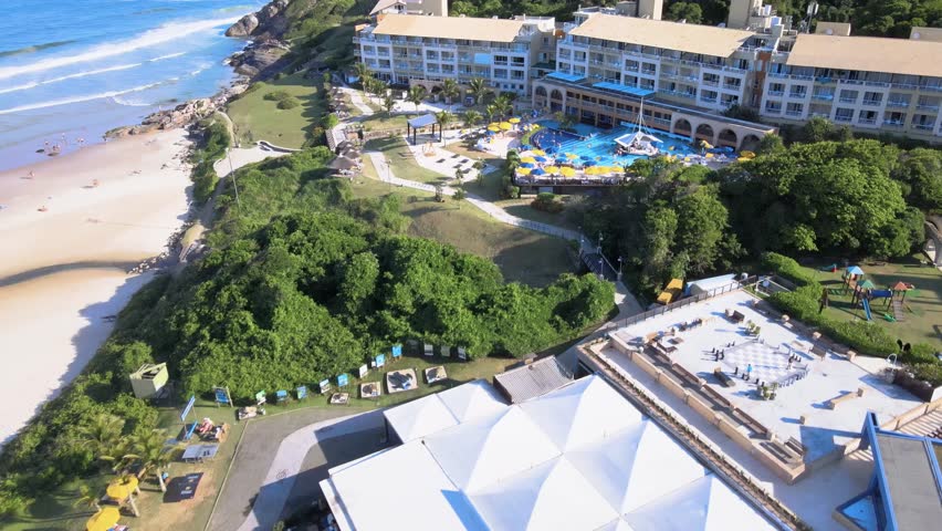High angle aerial view of the luxury Costao do Santinho Resort in Florianopolis, Brazil, featuring hotel buildings, swimming pools and rocky cliffs by the ocean waves on a sunny day.