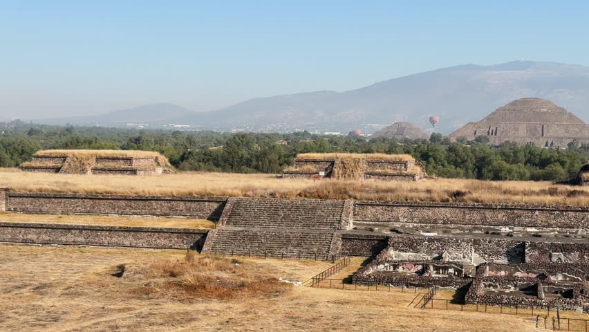 Scenic view across Teotihuacan ruins and terraces with dry grass foreground, distant pyramids, mountains, and hot air balloons—Mexico travel landscape b-roll.