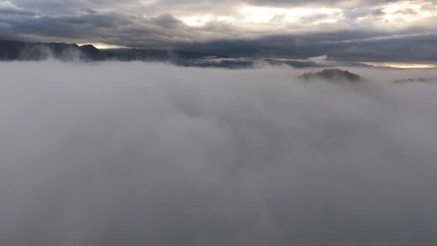 Aerial view rising over fog, dramatic cloudy sunset sky, in middle of alpines