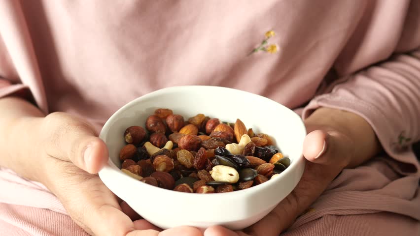 Close up video of a person holding a white bowl filled with a variety of healthy trail mix, picking up a cashew nut