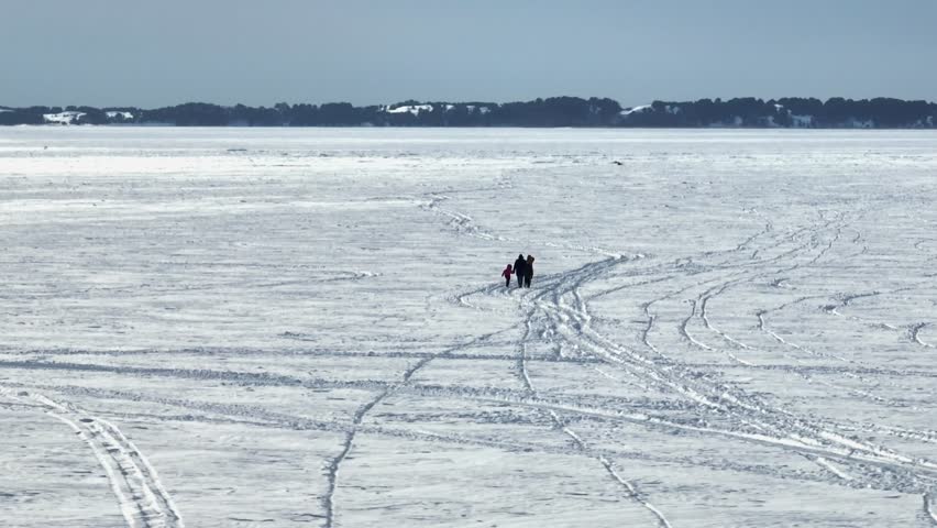 A distant, ground-level view of a small group trekking across a massive, snow-dusted frozen lake. Numerous tracks in the ice lead toward a dark, forested horizon under a hazy winter sky.