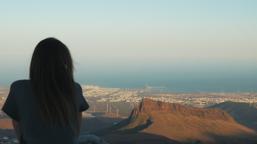 Back view of a woman sitting on a mountain peak. The girl is contemplating the beautiful landscape with a coastal city and sea. Gran Canaria.