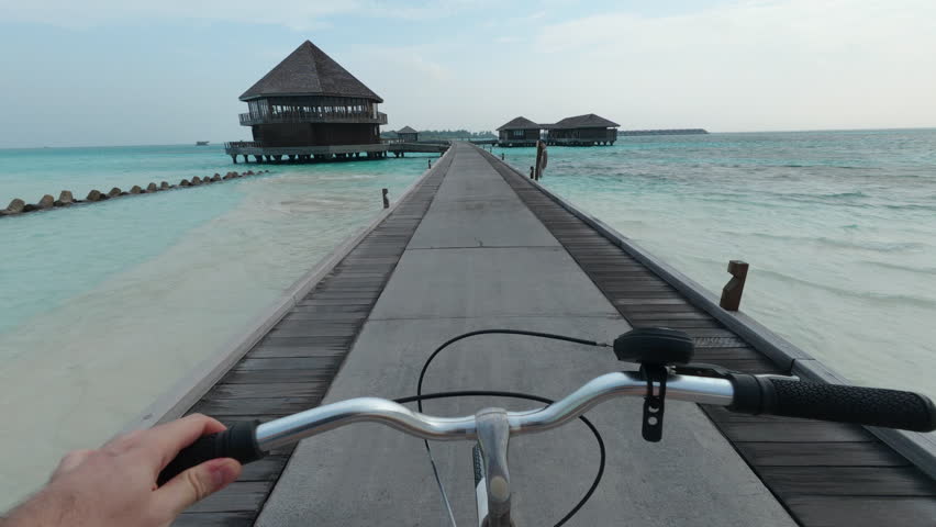 POV shot of a person riding a bicycle on a wooden bridge over the beautiful turquoise ocean passing overwater villas at a tropical Maldives resort under a partly cloudy sky