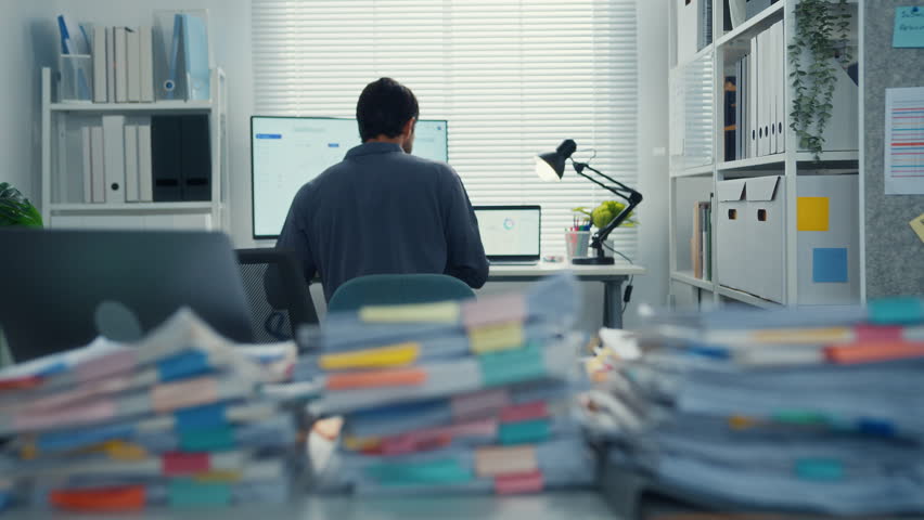 Overworked young Latin businessman reading document in large stack of paperwork files at office desk. Tax audit deadline, accounting stress, corporate burnout, bookkeeping, bureaucracy concept.