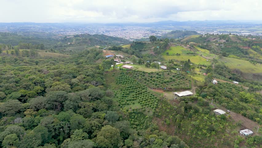 This aerial frame shows vibrant green fields surrounded by dense trees with a distant view of urban Popayán under a cloudy sky.