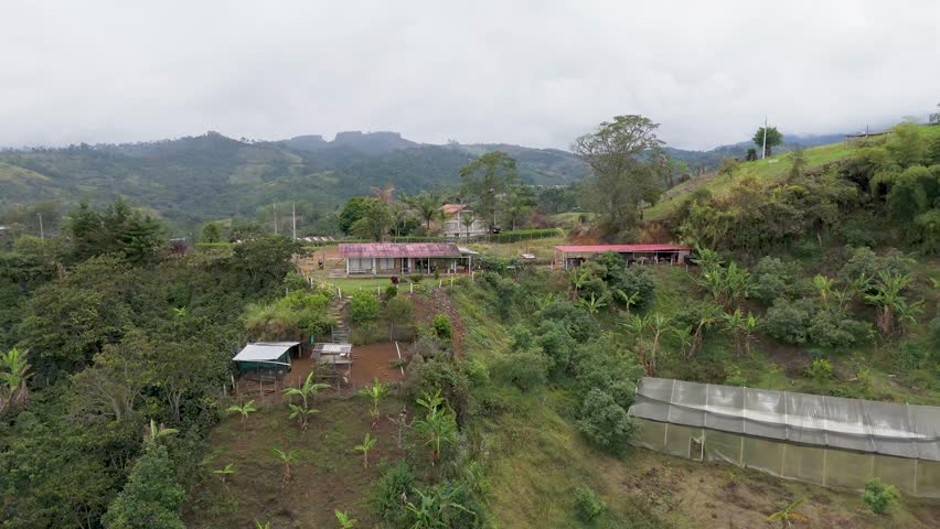 This aerial shot captures a green hilly landscape featuring several buildings and clear paths under a cloudy sky.