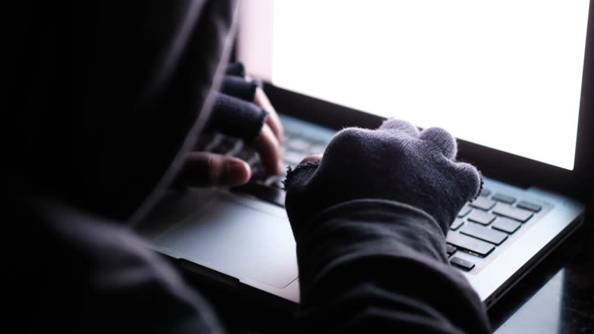 A close-up view of a person wearing a hooded sweatshirt and fingerless gloves typing on a laptop computer in a dimly lit room with glowing screen, dynamic shot