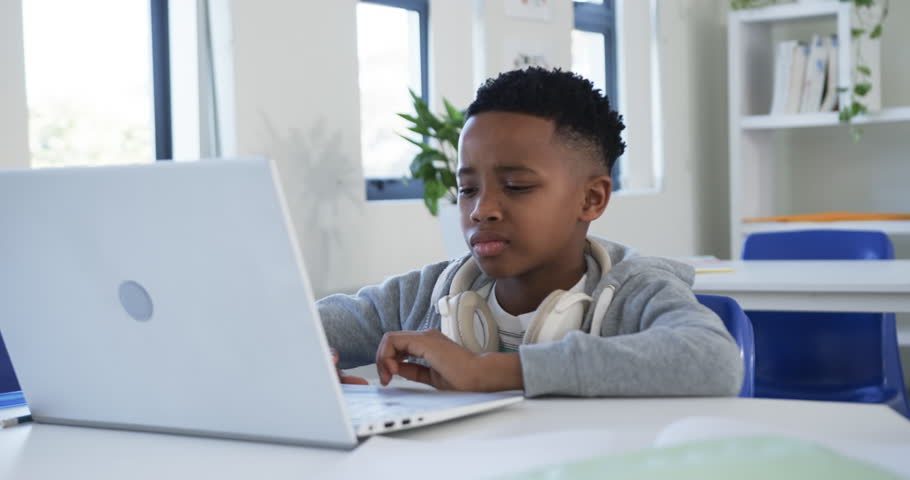 African male child typing on laptop at school desk after assignment alert, wearing white headphones. Student, computer, focus, classroom, learning, daylight, study