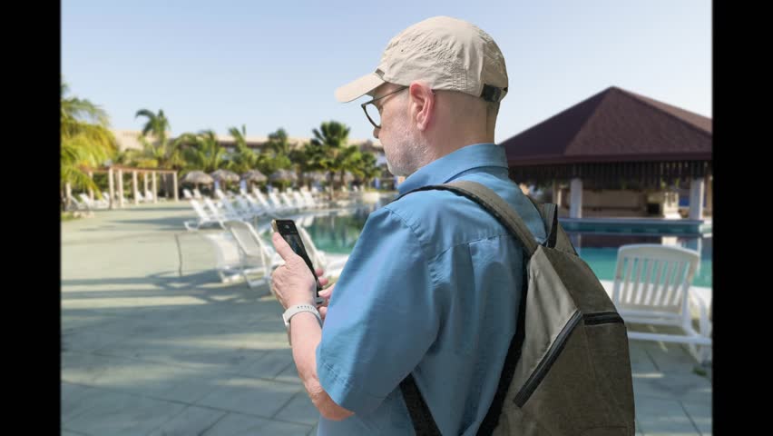 A man in a light cap and blue shirt checks his smartphone by a resort pool. A backpack rests on his back as palm trees and lounge chairs frame a tropical scene