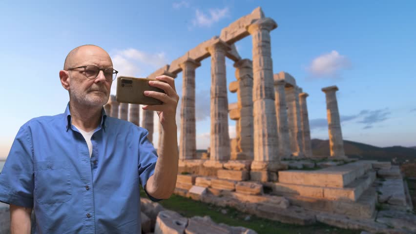 An older man in a blue shirt takes a selfie with ancient stone columns in the background. The sunset highlights the ruins, conveying travel, exploration, history, and personal moments.