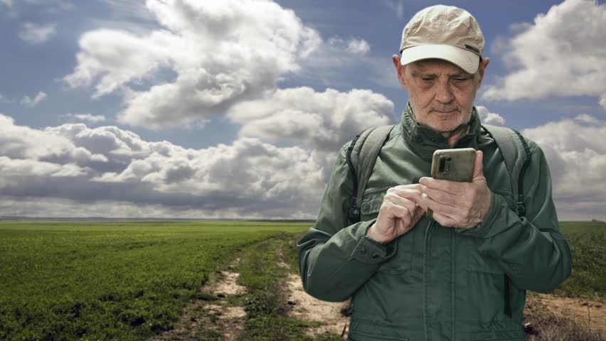 An older man in a green field, wearing a cap and backpack, stands on a dirt trail under a dramatic cloudy sky. A calm, outdoorsy moment capturing hiking, travel, and rural life.