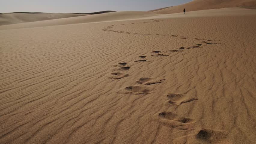 Wide horizontal view of human footprints trailing across rippled desert sand toward man figure walking towards distant dunes under clear sky