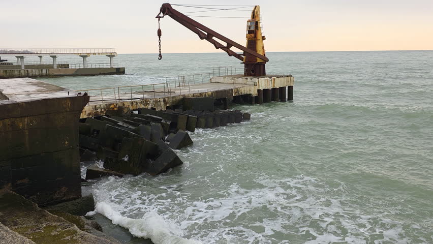 Old rusty industrial crane on a concrete pier with ocean waves crashing against the breakwater on a cloudy day, showcasing a tranquil yet powerful coastal scene