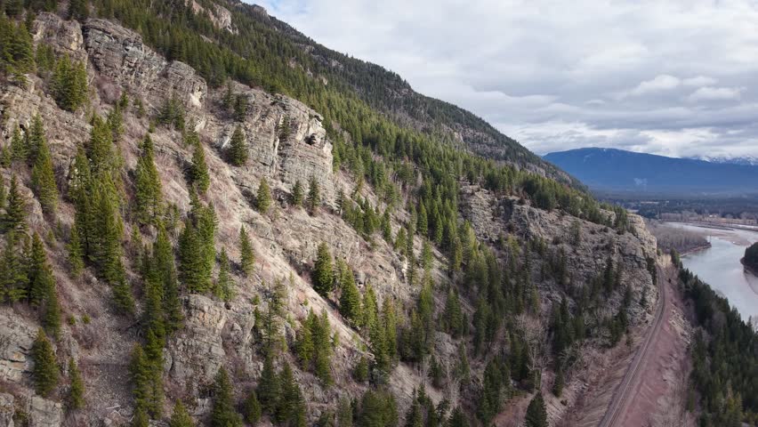Aerial drone footage of a rocky forested hill dropping down to a winding river and road near Whitefish, Montana, with mountains and sky in the background