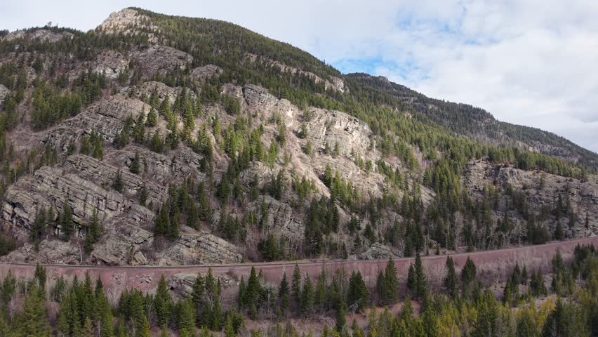 Aerial drone view of a forested rocky hillside and mountain slope with evergreen trees and a road at the base near the Flathead River valley in Whitefish, Montana.