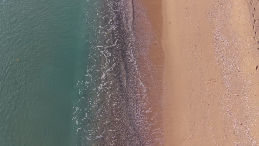 Top view aerial of green sea meeting sandy beach shoreline with gentle waves creating natural abstract coastal pattern.