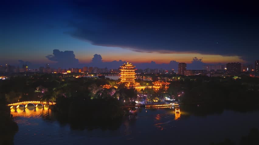 Aerial view of Jinan, China, at dusk. Chaoran Tower glows golden by Daming Lake, lights reflecting on calm water. Vibrant city skyline frames this serene urban night scene.