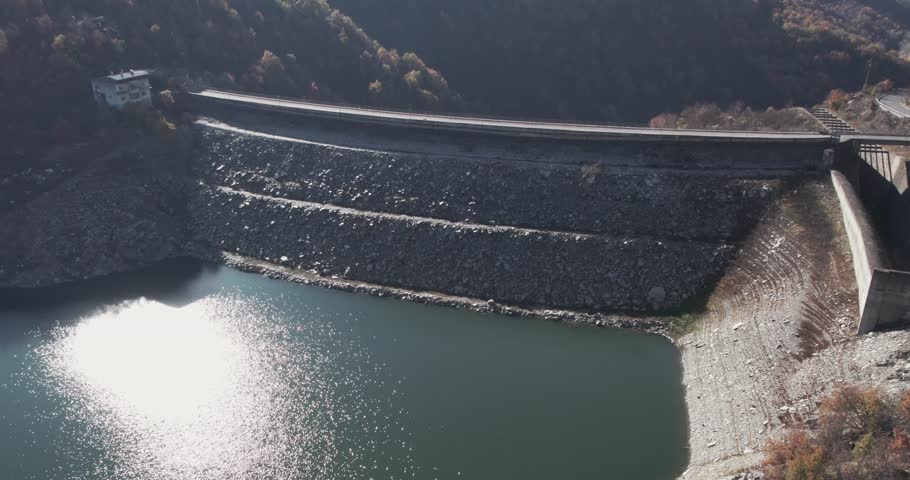 Empty concrete mountain dam bridge surrounded by lush tree-covered hills during drought season