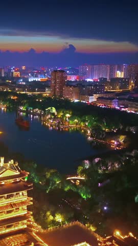 Stunning aerial view of Jinan City at dusk, showcasing the beautifully lit Chaoran Tower by Daming Lake. The vibrant city lights illuminate the traditional architecture, scenic lake, and modern skyline under a colorful twilight sky. A captivating blend of history and urban life.