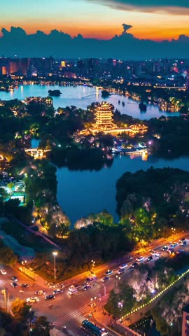 An aerial shot captures the beautifully illuminated Chaoran Tower on Daming Lake in Jinan City, China, at twilight. The vibrant lights reflect on the calm water, surrounded by lush trees and city lights in the distance, creating a serene and picturesque evening scene.