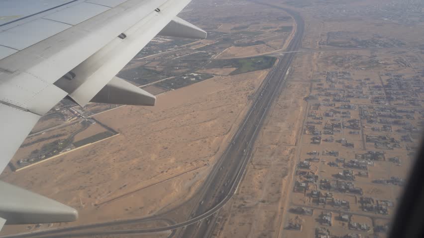Airplane wing extends over a vast desert landscape, revealing a network of roads and patches of green fields, showcasing the contrast between urban development and natural terrain