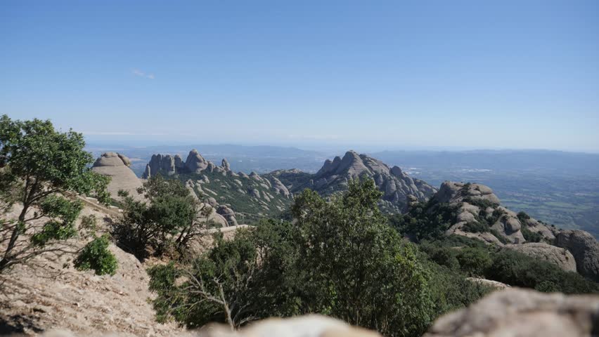 A wide-angle timelapse showing hikers ascending and descending the rugged, conglomerate rock trails of Montserrat under a bright blue sky in Catalonia.