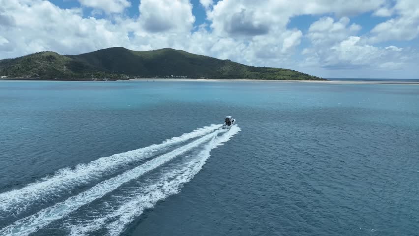 Slow drone tracking shot in daytime, following behind a speedboat racing across turquoise Whitsundays water toward a sandy shoreline and green island hills, Queensland, Australia.