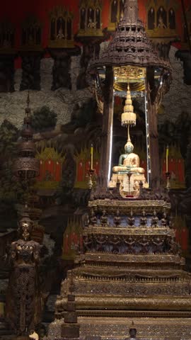 Jade Buddha rests on an ornate altar inside Wat Phra Kaew or Temple of the Emerald Buddha in Bangkok, vertical handheld right pan.