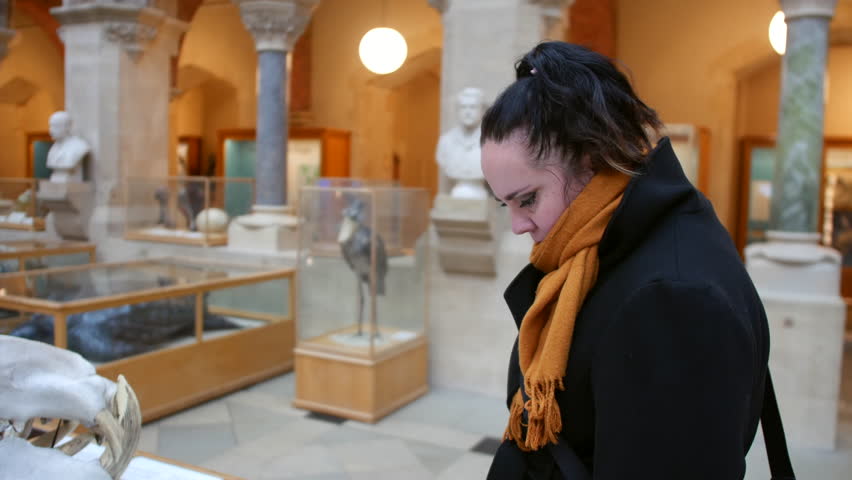 Woman observing museum exhibit while standing in historic gallery with glass display cases and classical busts. Visitor wearing black coat and mustard scarf examining specimen inside natural history m