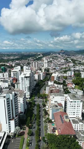 Vertical Aerial View of Modern City Skyline with High-Rise Buildings, Tennis Courts, and Lush Green Hills in Background