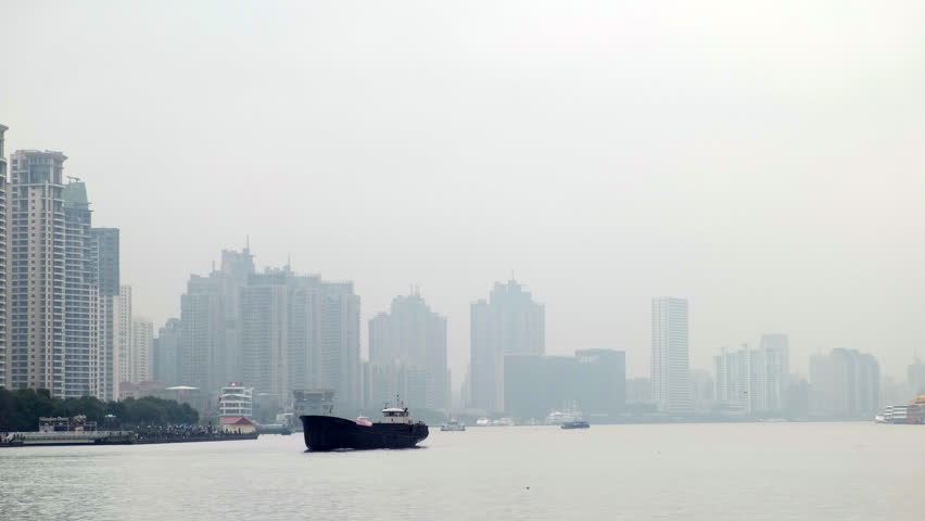 Boats navigate a wide river in front of the hazy, heavily polluted urban skyline of Shanghai China on an overcast day.