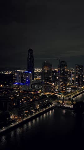 Vertical aerial pan right of Puerto Madero high rise buildings and waterfront at night in Buenos Aires. Bright city lights and river reflections.