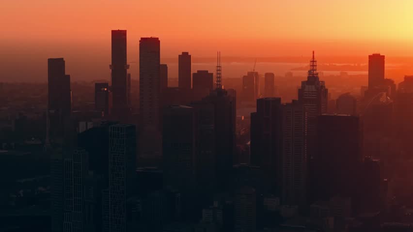 Deep orange dusk over Melbourne CBD with darkened skyscraper silhouettes and fading daylight across the skyline, aerial medium ascend overview