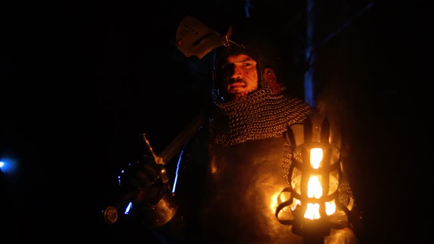 A knight in full armor, holding a sword and torch, walks through a dark night forest. A medieval warrior searches in the darkness.