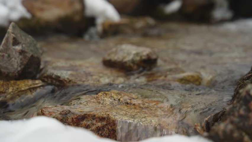 Clear mountain stream flowing over granite stones with snow on the banks. Serene nature scene showing a close up of a cold, rocky riverbed, capturing the peaceful movement of fresh water