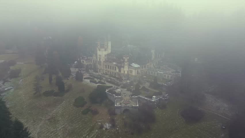 High quality cinematic drone reveal of the historic Peles Castle through thick mountain mist in Sinaia Romania featuring stunning Neo Renaissance architecture and a mysterious moody atmosphere