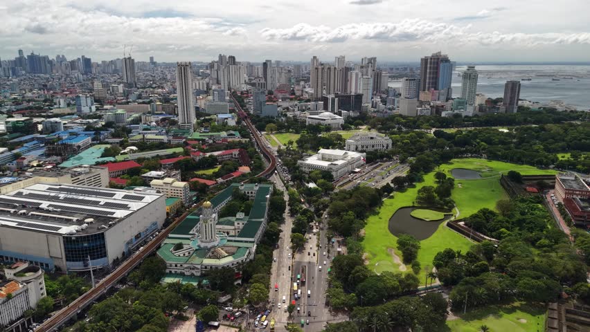Wide aerial panorama over Rizal Park reveals green open spaces and ornamental pond with classical government buildings and dense Manila skyline stretching toward Manila Bay under overcast sky