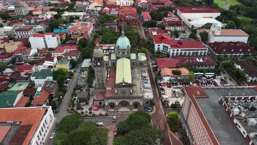 Elevated frontal aerial shows full length of Manila Cathedral from stone facade and plaza entrance through nave to central dome flanked by Intramuros colonial streets with terracotta rooftops