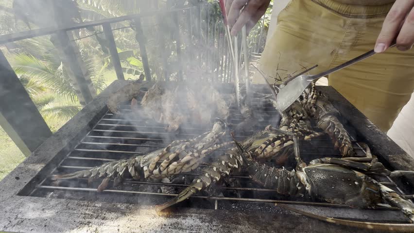 Chef turning whole lobsters with tongs and spatula on charcoal grill over open fire in tropical outdoor setting.
